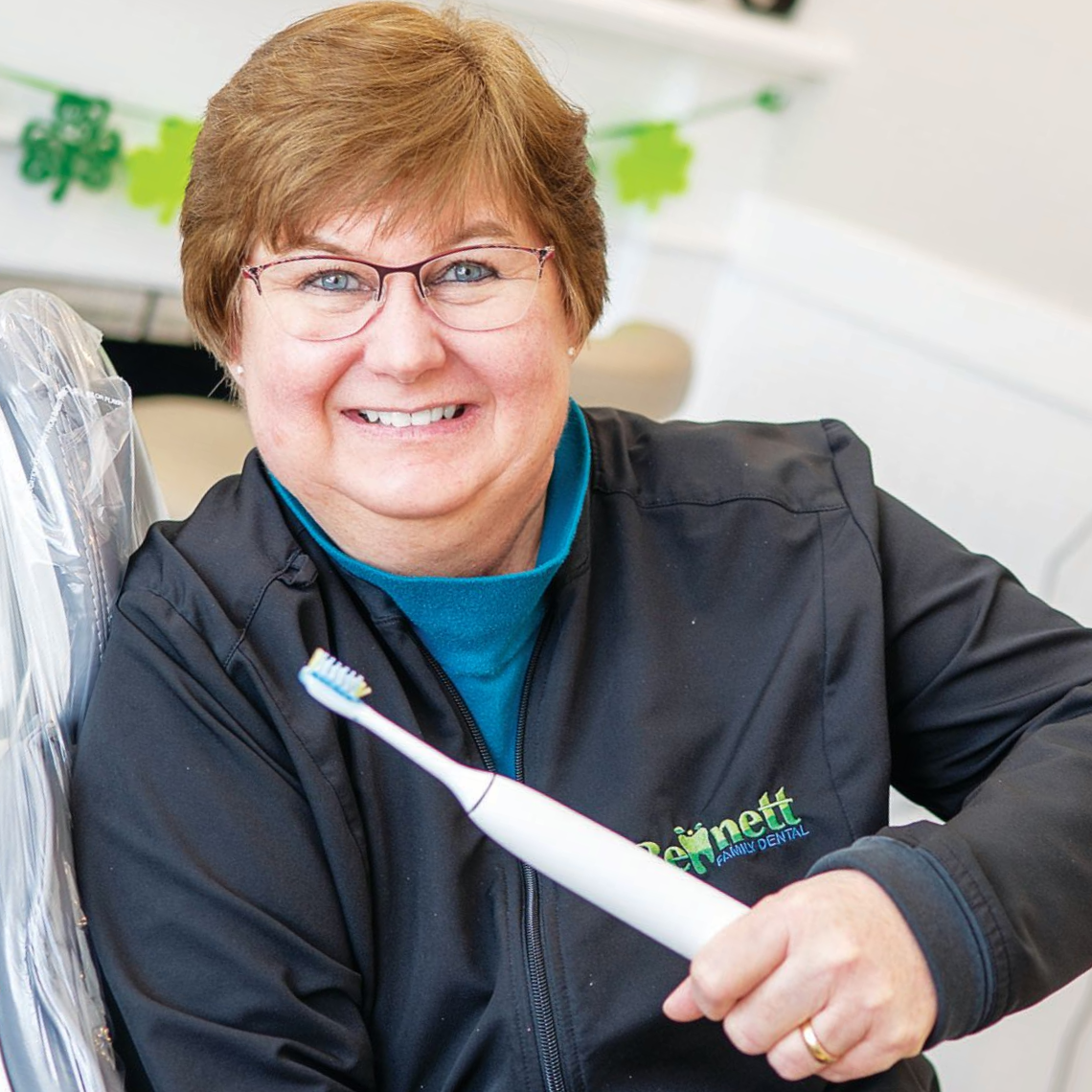 A smiling person in a teal top and black jacket holds an electric toothbrush in an office with shamrock decorations.
