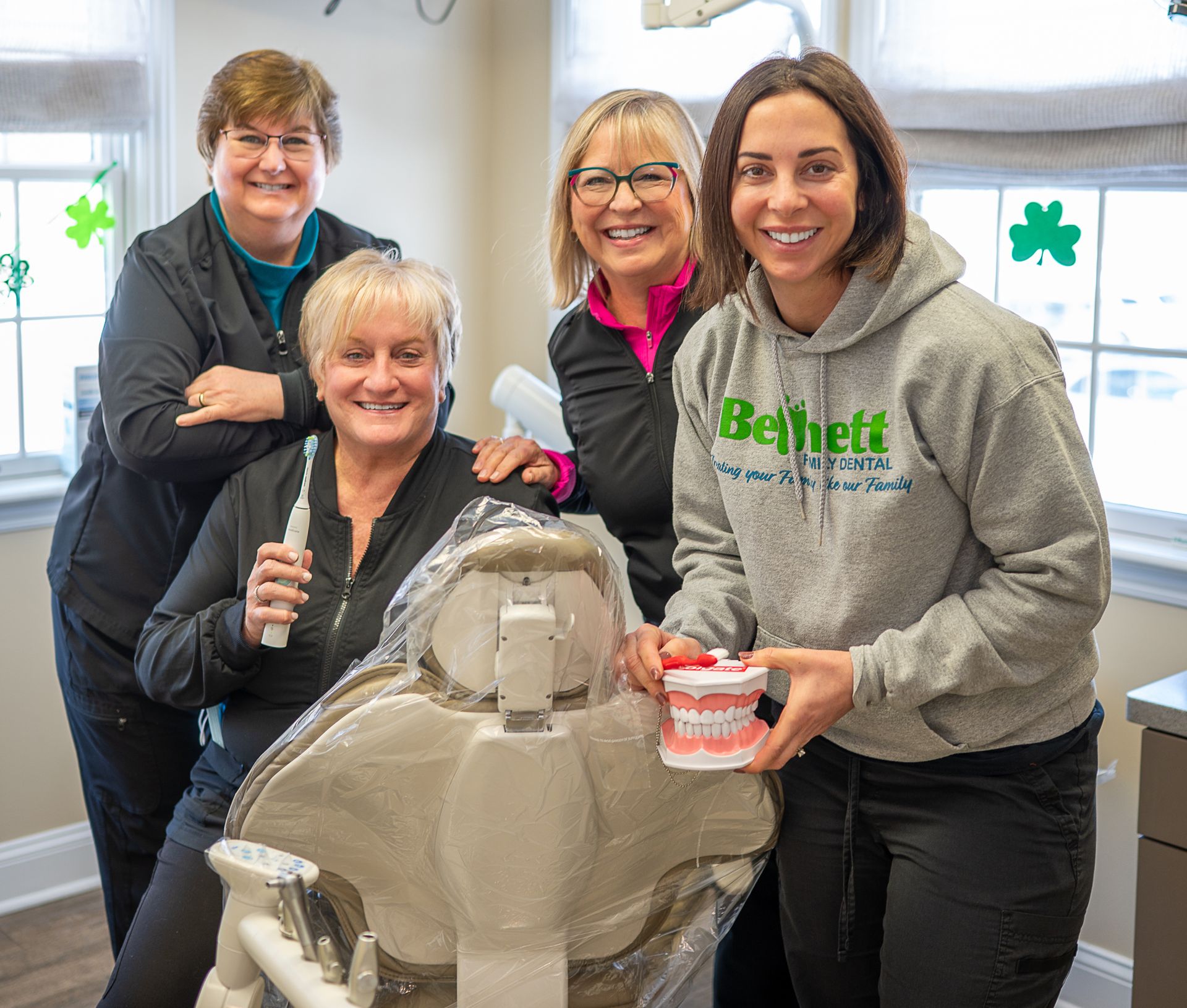 Four smiling dental staff members stand in an office, one holding a dental model and another holding an electric toothbrush.