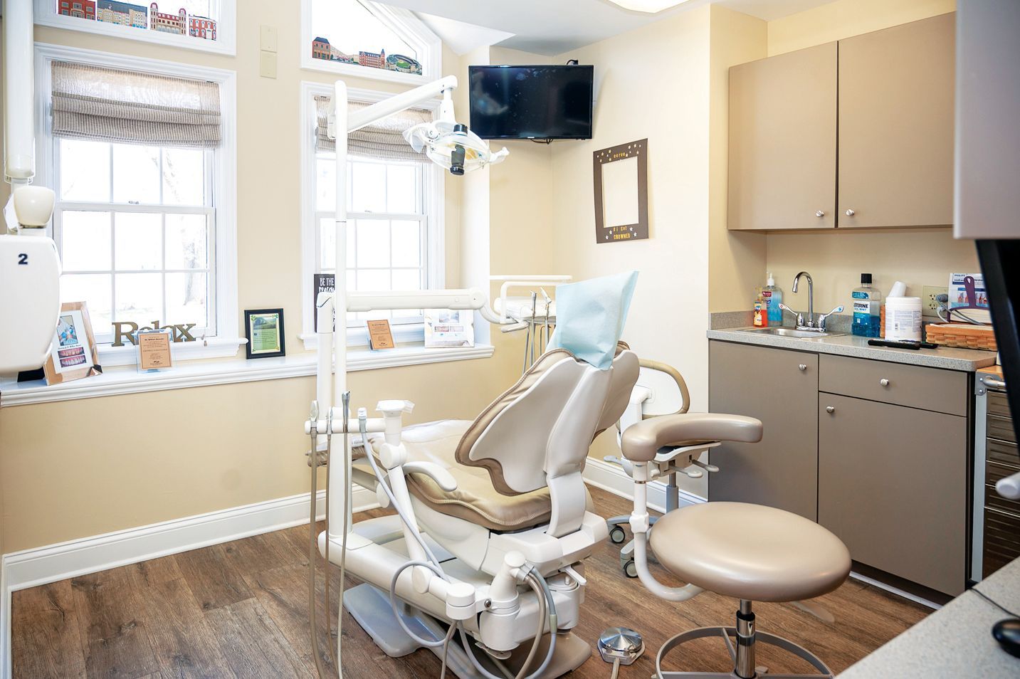A dental examination room with a beige treatment chair, a practitioner’s stool, cabinetry, and a window with blinds.