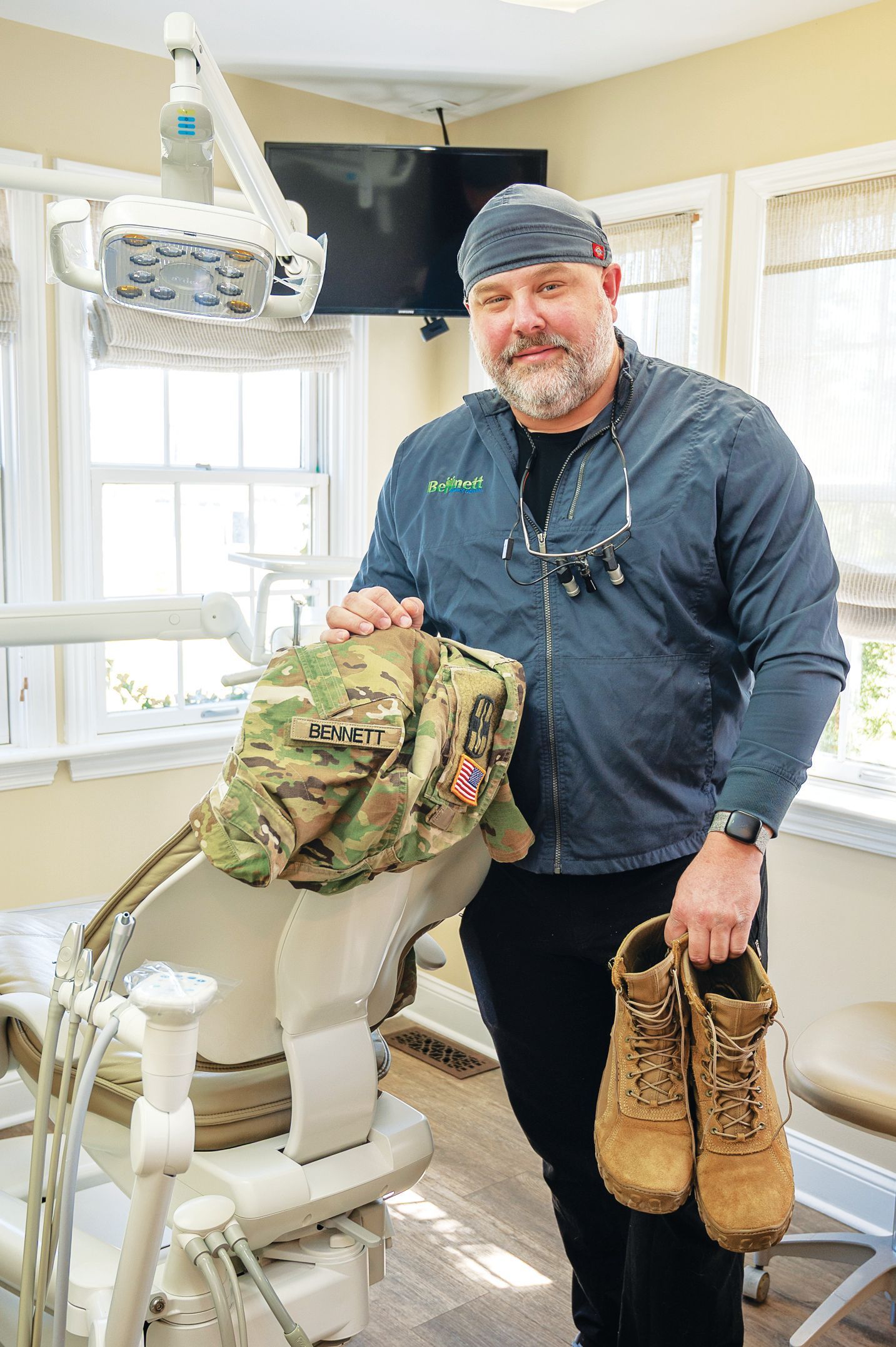 A person wearing a surgical cap stands in a dental office holding military combat boots and a camouflage backpack.
