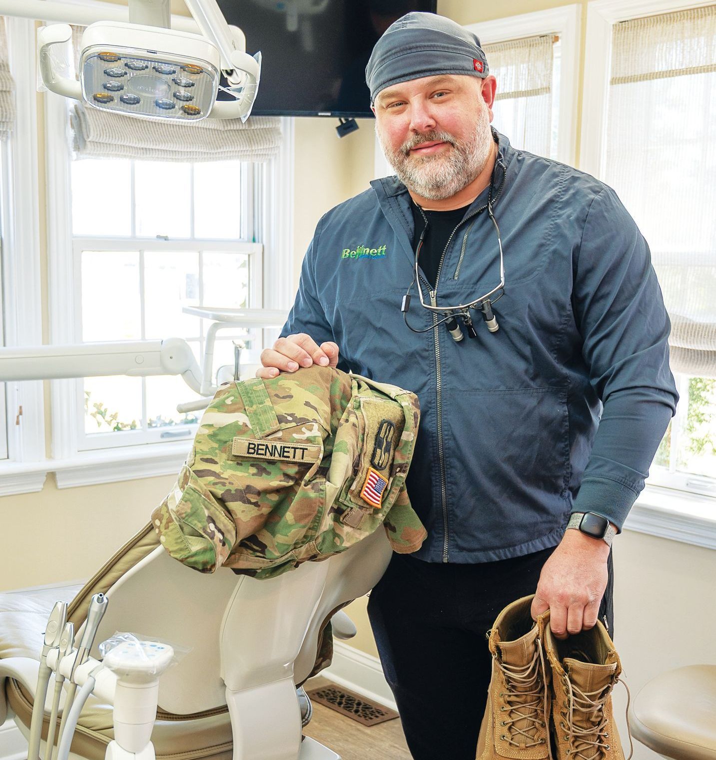 A man in a dental office holds a camouflaged military uniform jacket and a pair of tan combat boots.