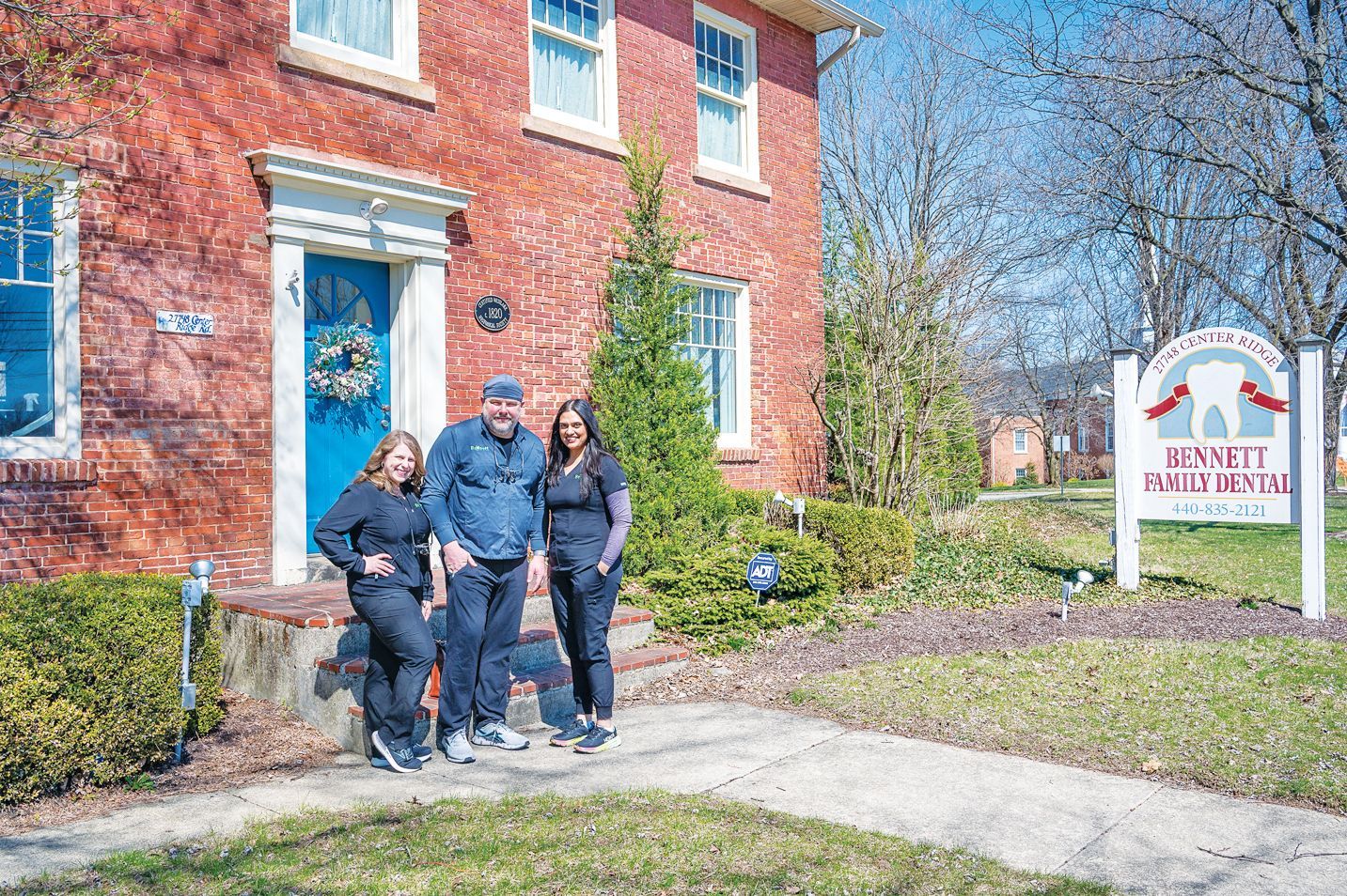 Three people standing in front of a brick building with a sign for Bennett Dentistry on a sunny day.
