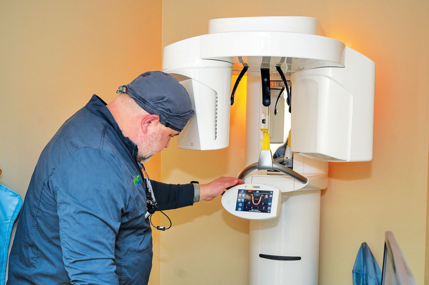 A professional wearing a scrub cap operates a dental panoramic X-ray machine in a clinical office setting.