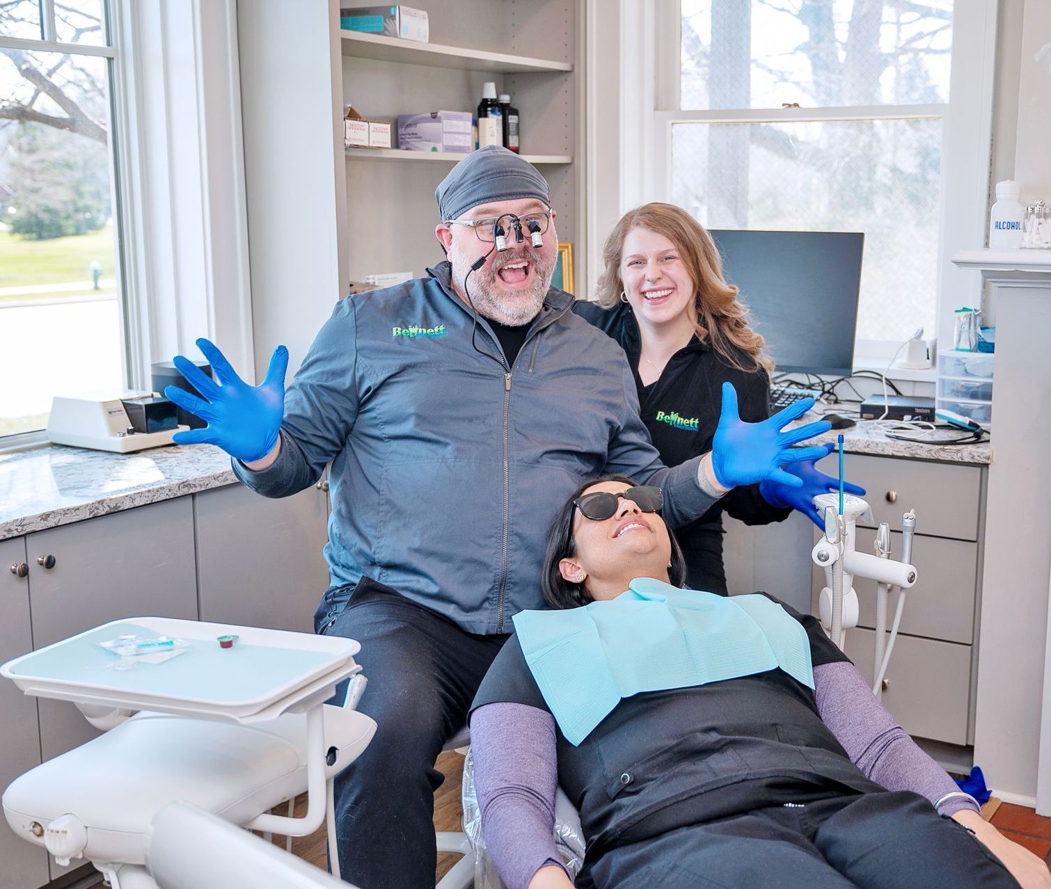A dental team in scrubs poses behind a patient in a dental chair; the dentist makes a silly face with hands raised.