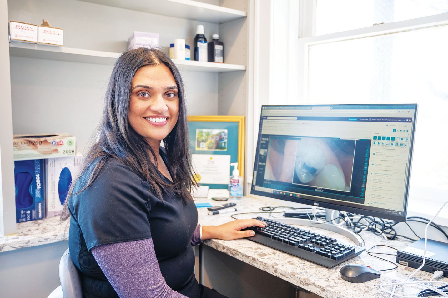 Professional working at a computer desk in a medical office setting, looking toward the camera.