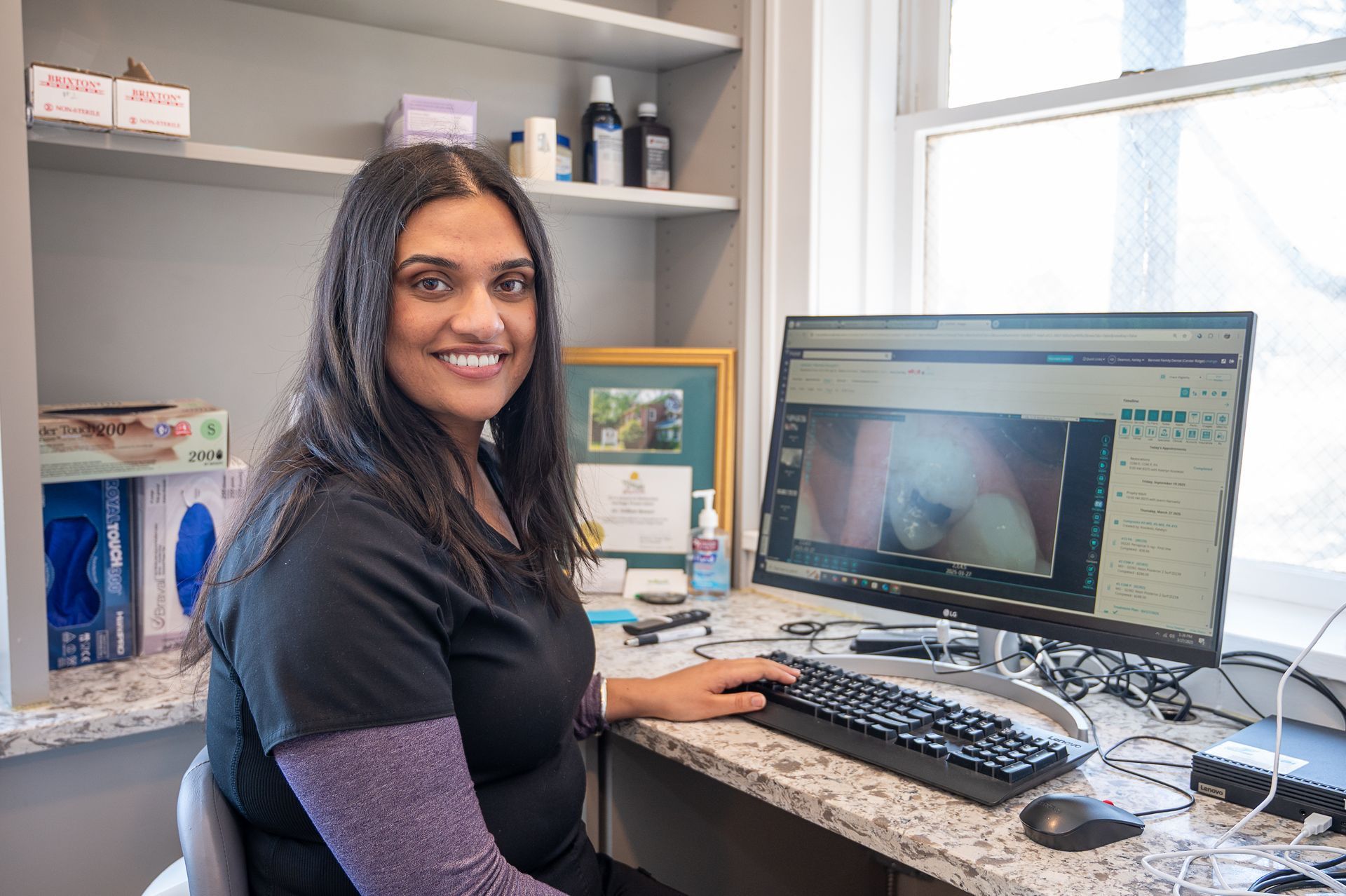 A smiling person in a black scrub top sits at a desk in an office, looking at a computer screen showing dental imaging.