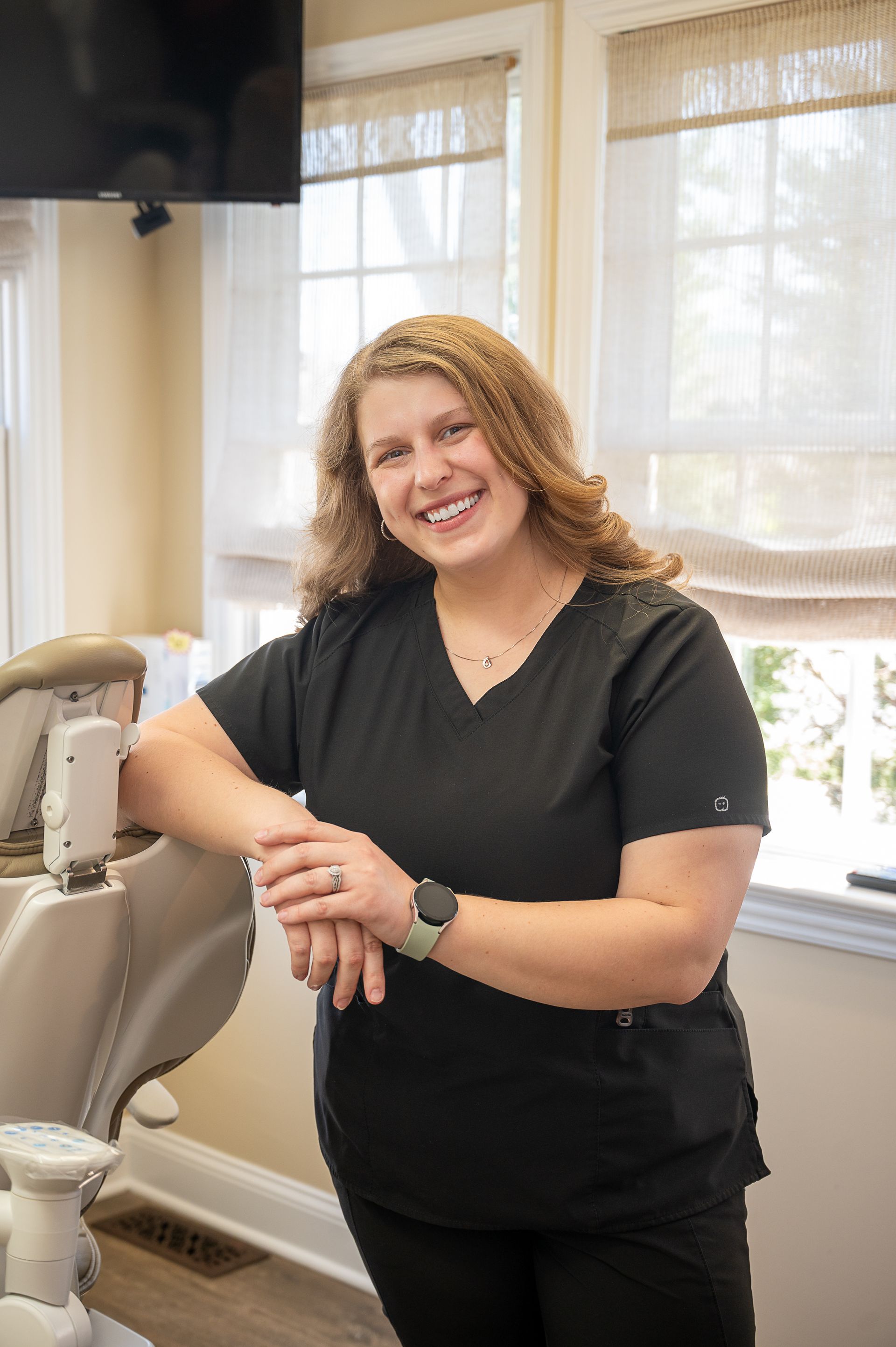 A person in black scrubs smiles while leaning against a dental chair in a bright office.