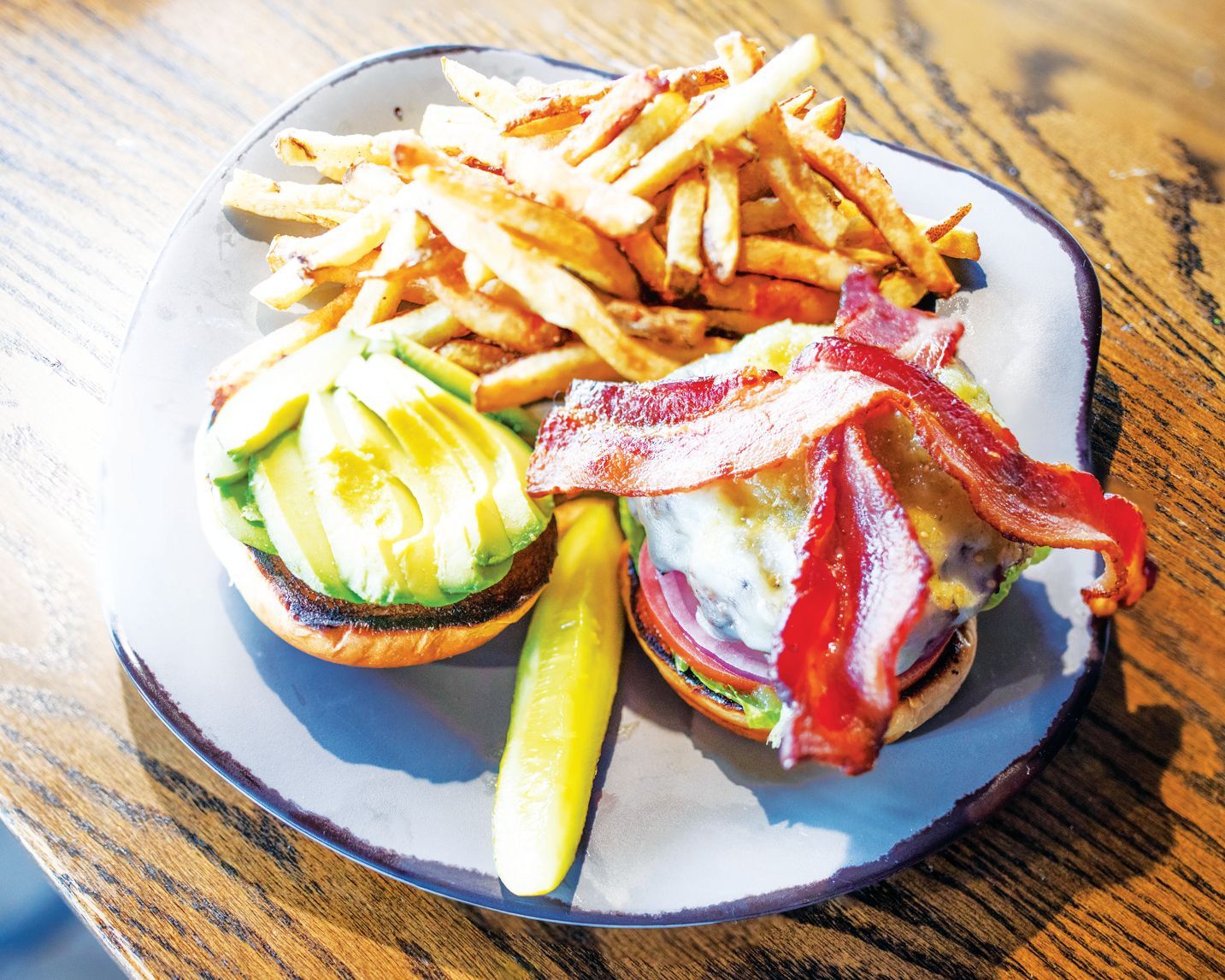 An open-faced burger with bacon, avocado, and a pickle, served with a side of french fries on a plate.