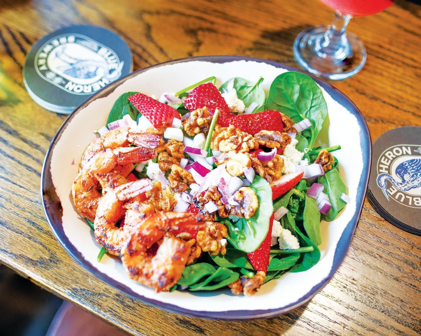 A bowl of spinach salad topped with grilled shrimp, sliced strawberries, crumbled cheese, and walnuts on a wooden table.