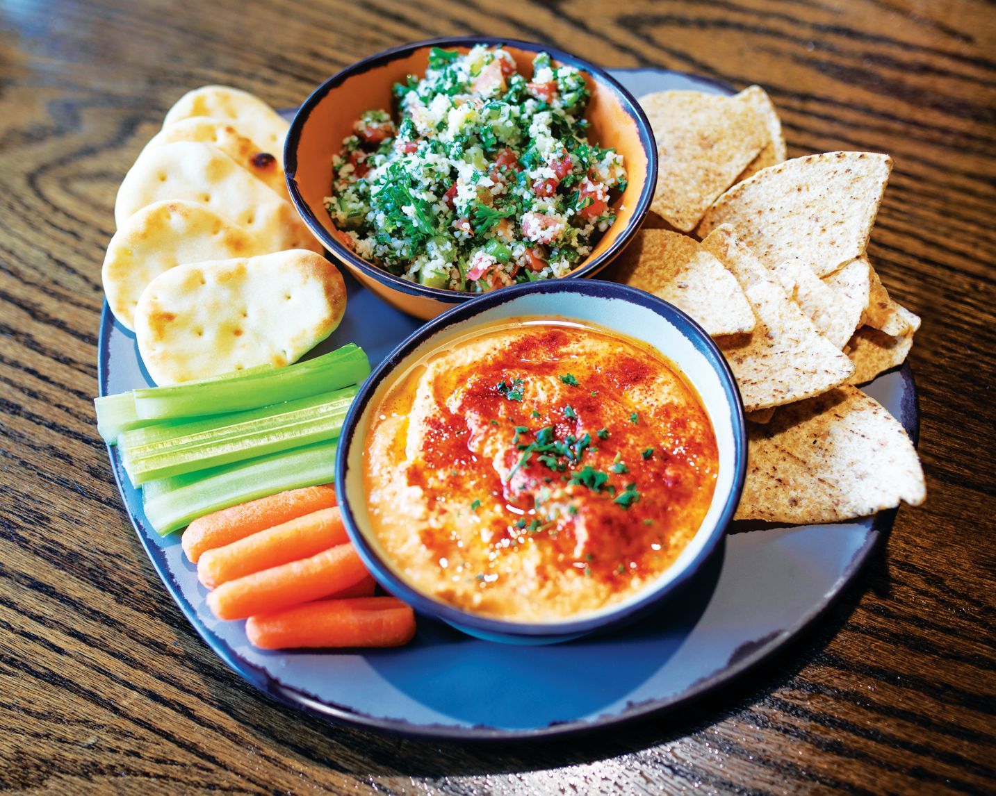 A platter with two bowls of dips, pita chips, crackers, celery sticks, and baby carrots on a dark wood table.