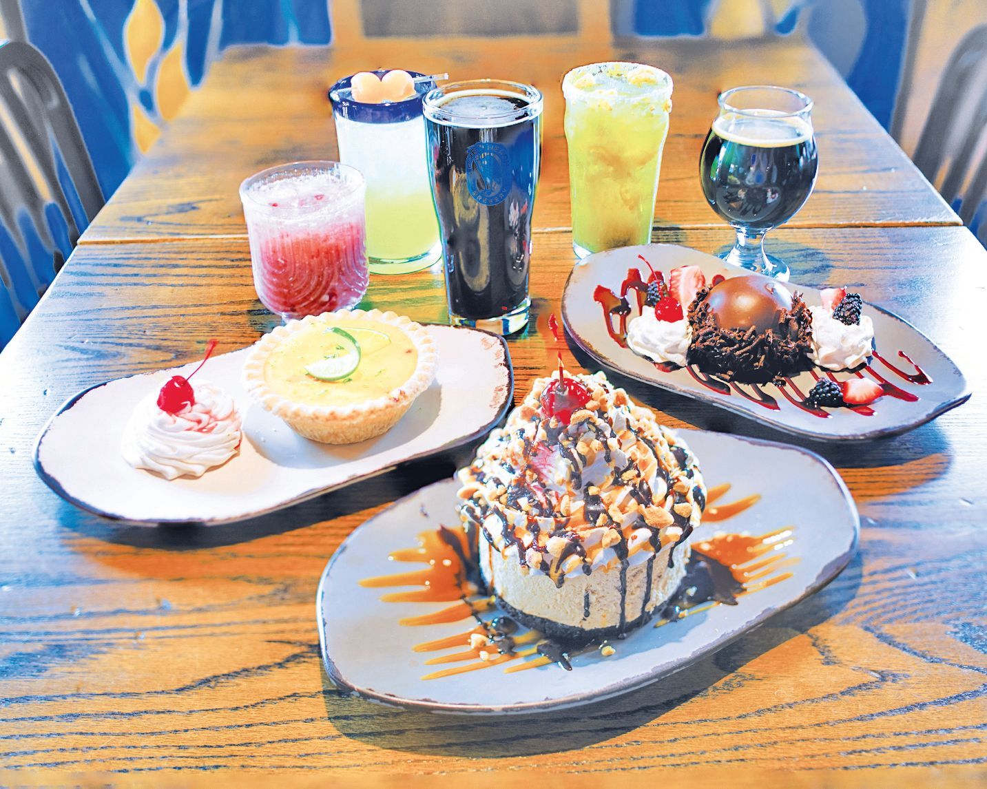A wooden table displaying an assortment of colorful cocktails and decadent desserts on ceramic plates.
