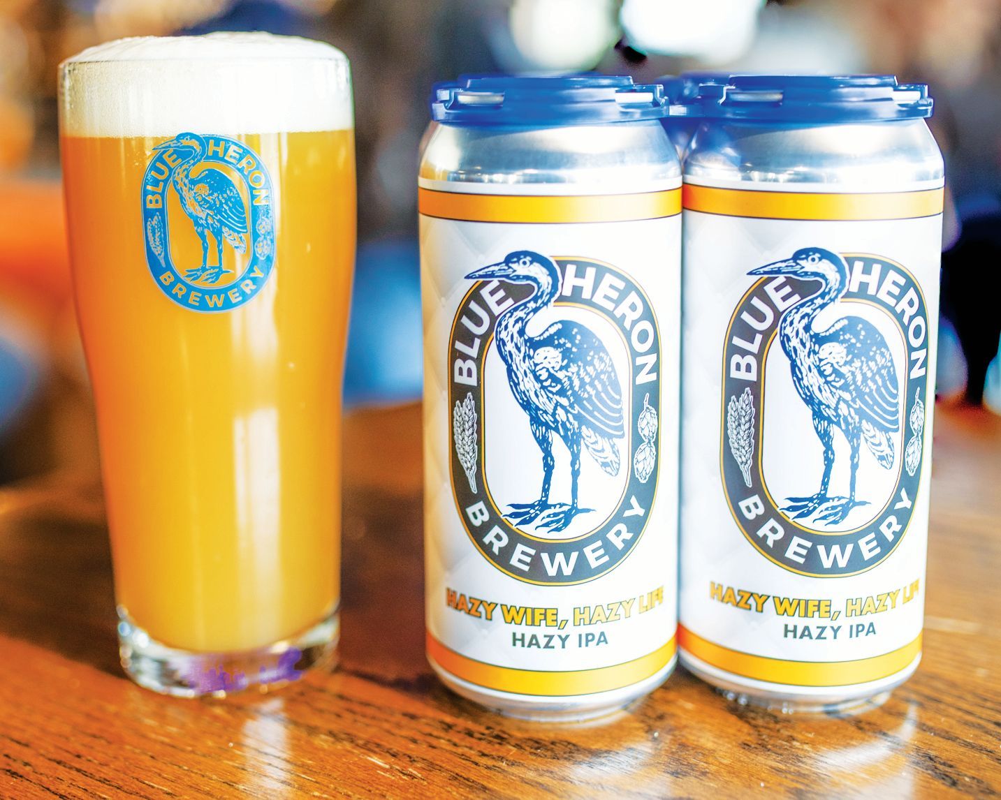 A glass of hazy golden beer next to two cans of Blue Heron Brewery Hazy IPA on a wooden table.