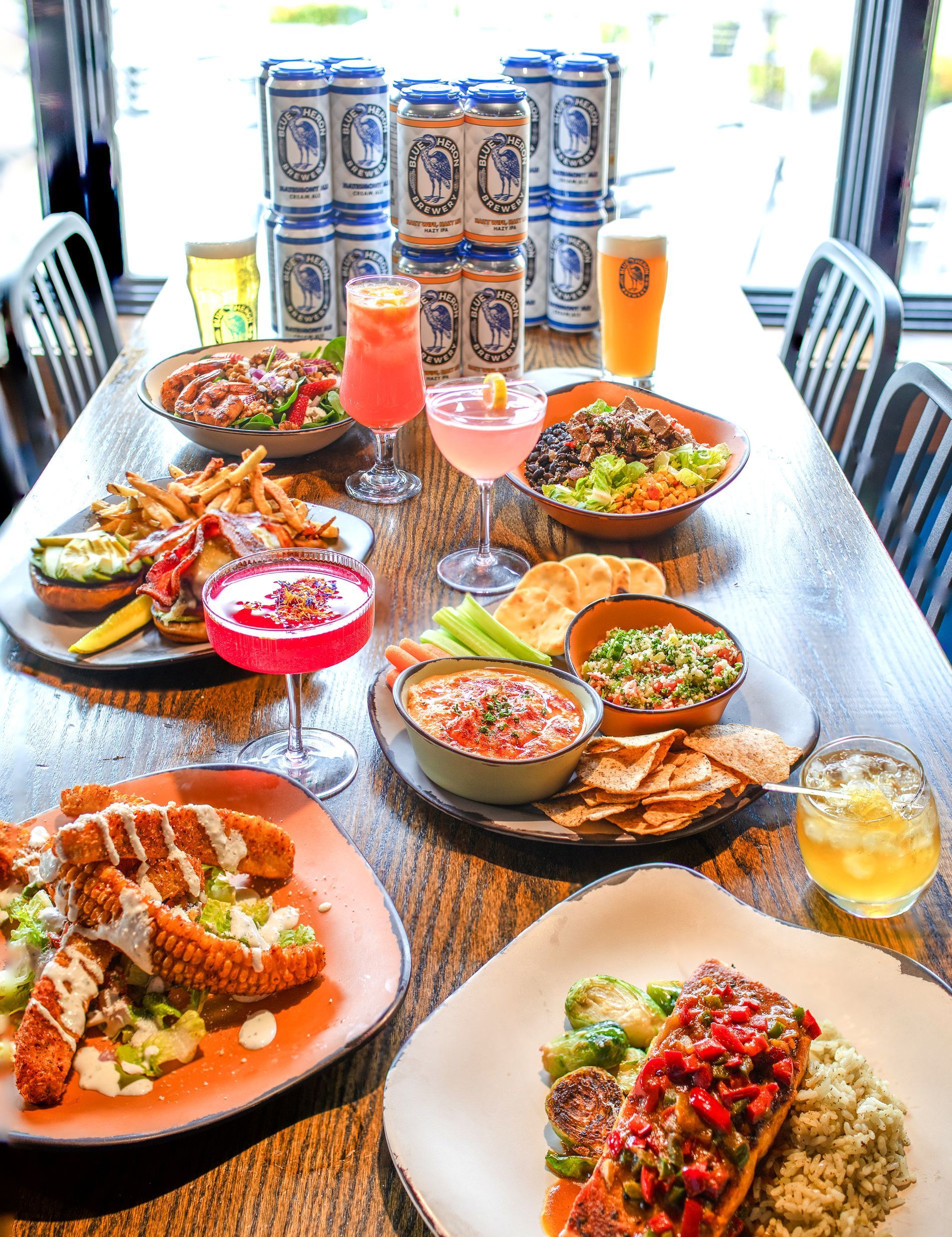 A table filled with various dishes, cocktails, and canned drinks in a restaurant setting.