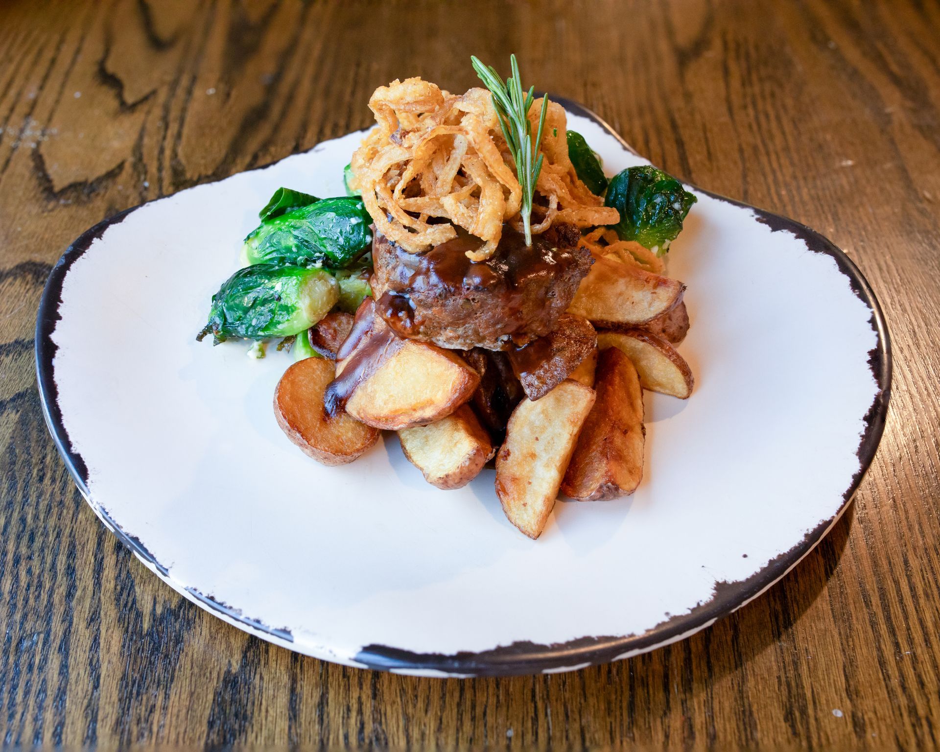 A serving of steak topped with crispy fried onions, served with potato wedges and greens on a white speckled plate.