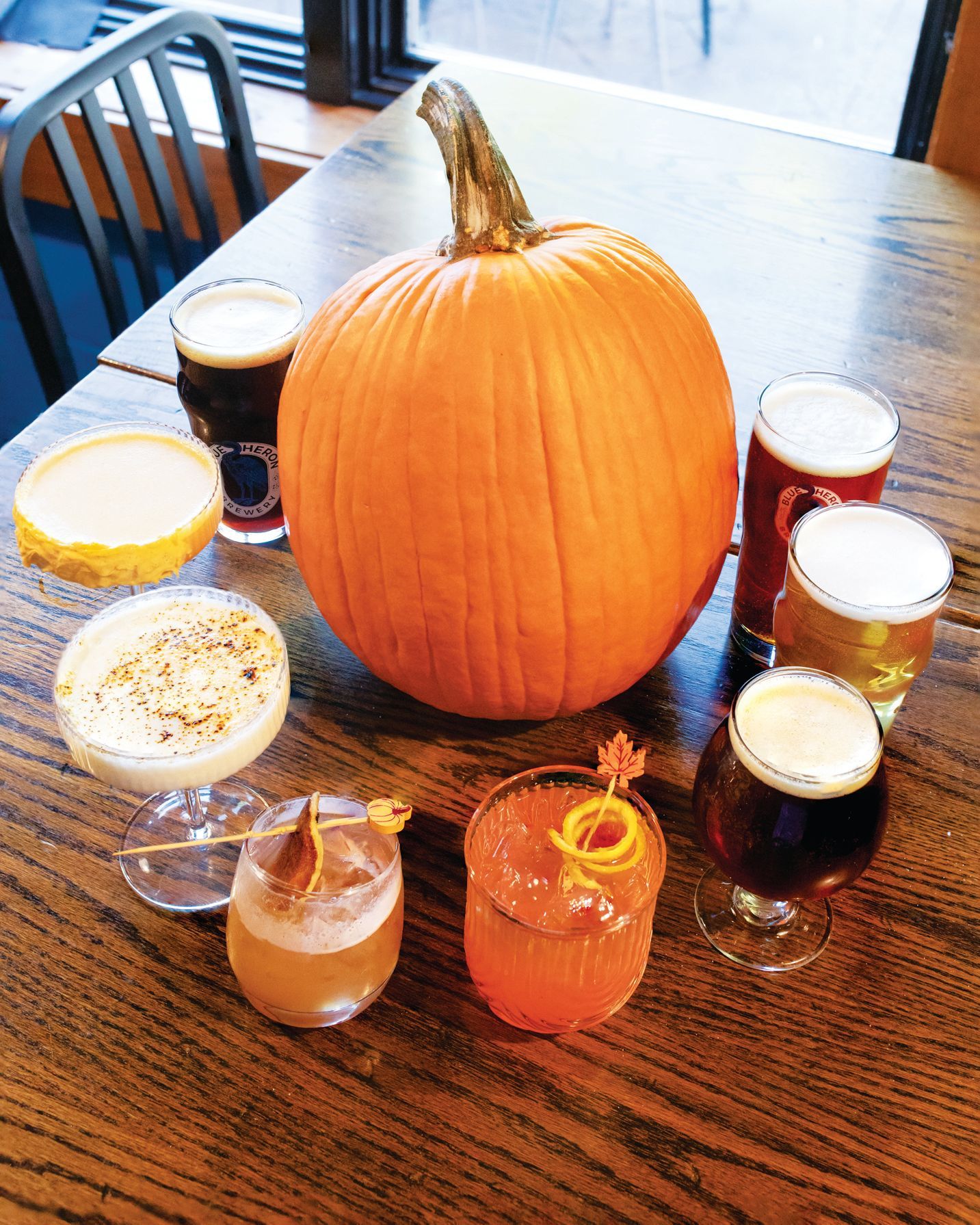 A wooden table featuring a large orange pumpkin surrounded by an assortment of cocktails and craft beers.