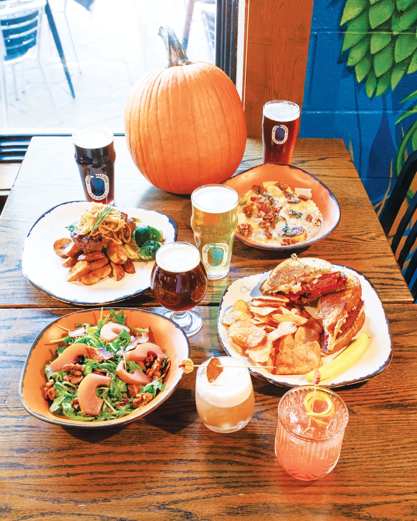 A wooden table laden with various seasonal dishes, cocktails, and beers, centered by a large orange pumpkin.