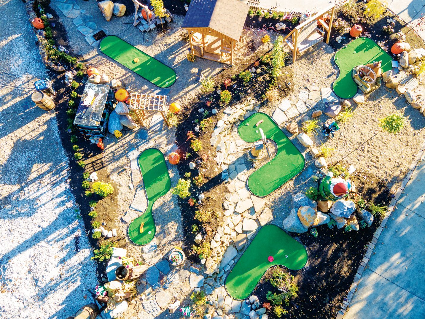 An aerial view of a mini-golf course with green putting patches, rock landscaping, and decorative pumpkins on sunny ground.