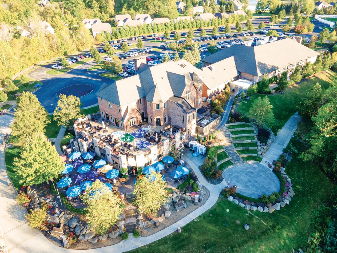 Aerial view of a multi-story brick venue with an outdoor patio filled with tables, blue umbrellas, and lush landscaping.