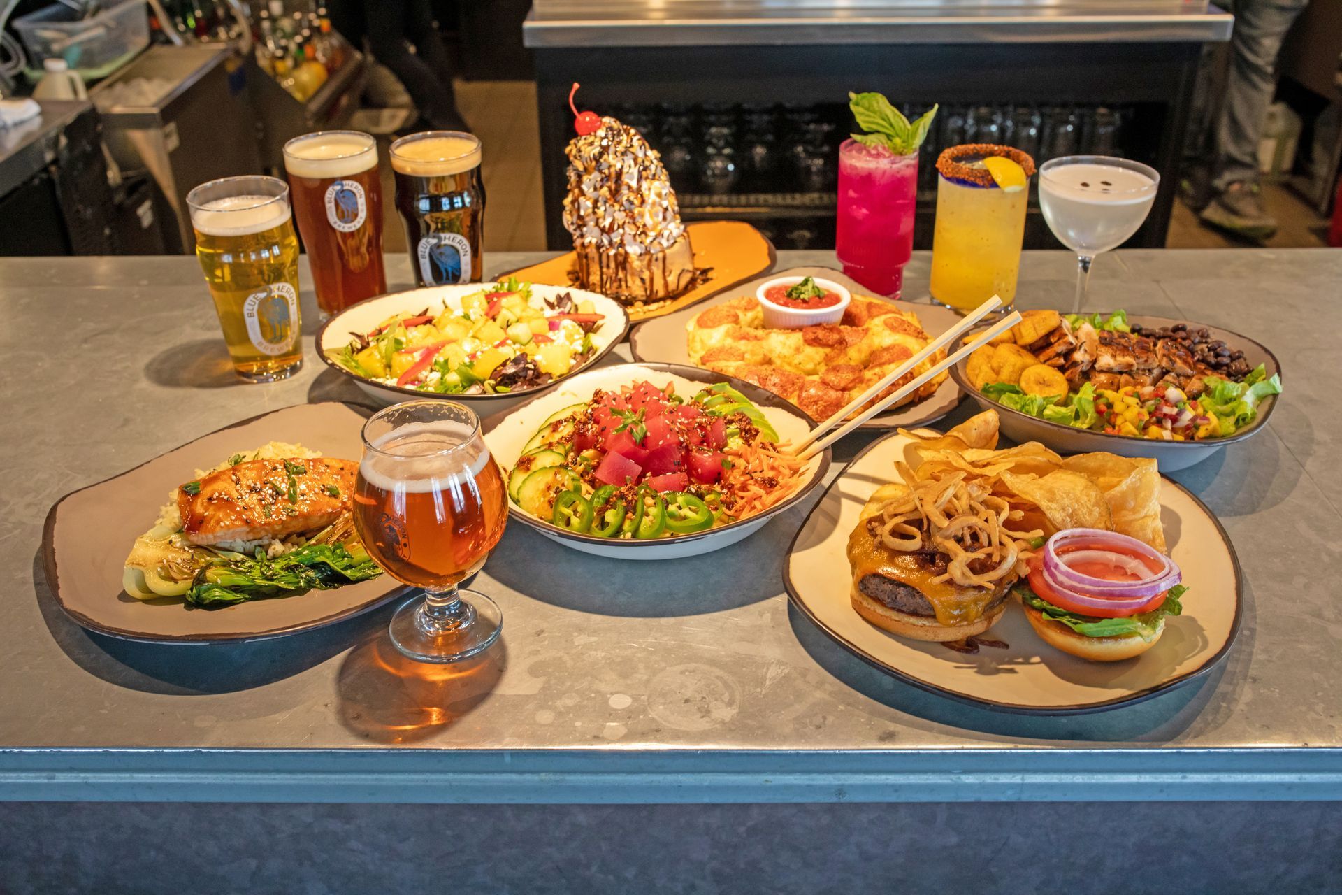 A spread of assorted appetizers, main dishes, and colorful cocktails displayed on a restaurant bar counter.