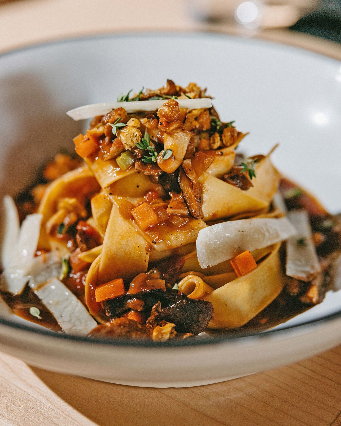A white bowl of wide, golden-brown pasta topped with sautéed mushrooms, diced carrots, herbs, and shavings of hard cheese.
