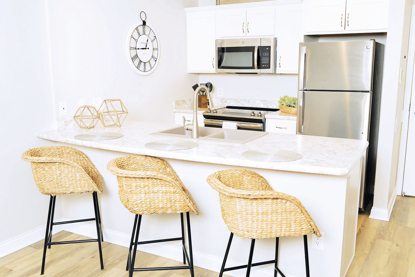 A modern kitchen with a white breakfast bar, three woven bar stools, stainless steel appliances, and a wall clock.