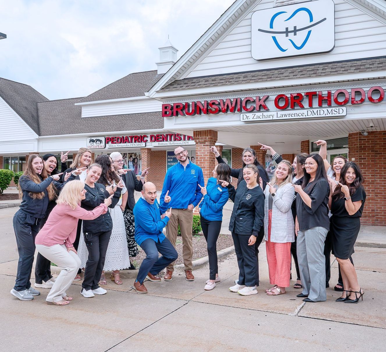 A large group of staff members in professional attire stand posing and smiling outside the Brunswick Orthodontics building.