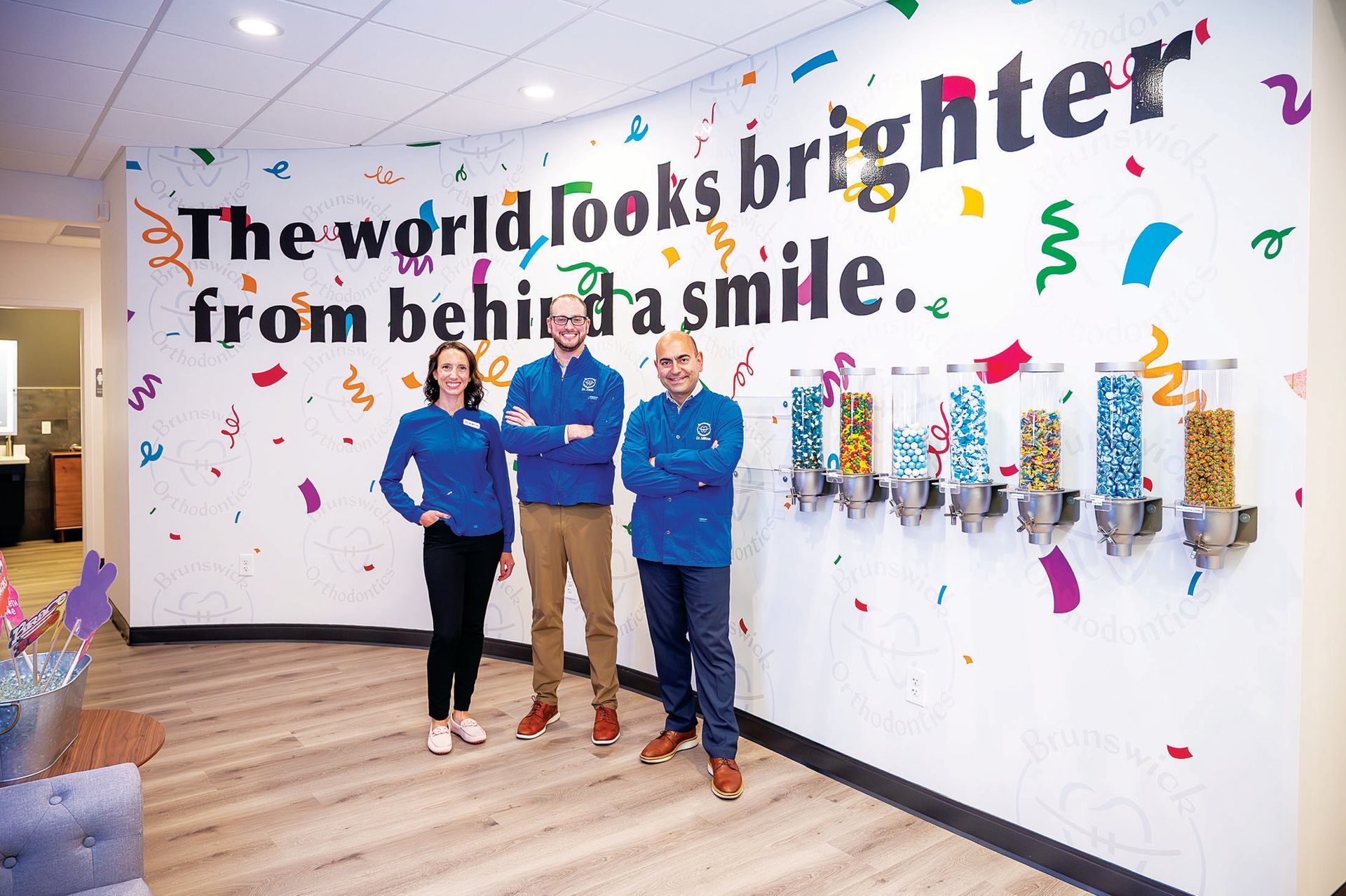 Three people stand smiling in front of a white wall decorated with confetti and the text 