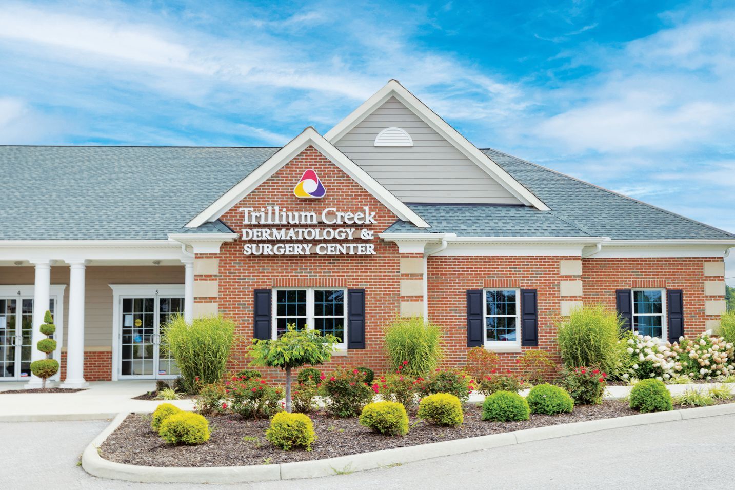 A brick, single-story Trillium Creek Dermatology and Surgery Center building with a landscaped front garden under a blue sky.
