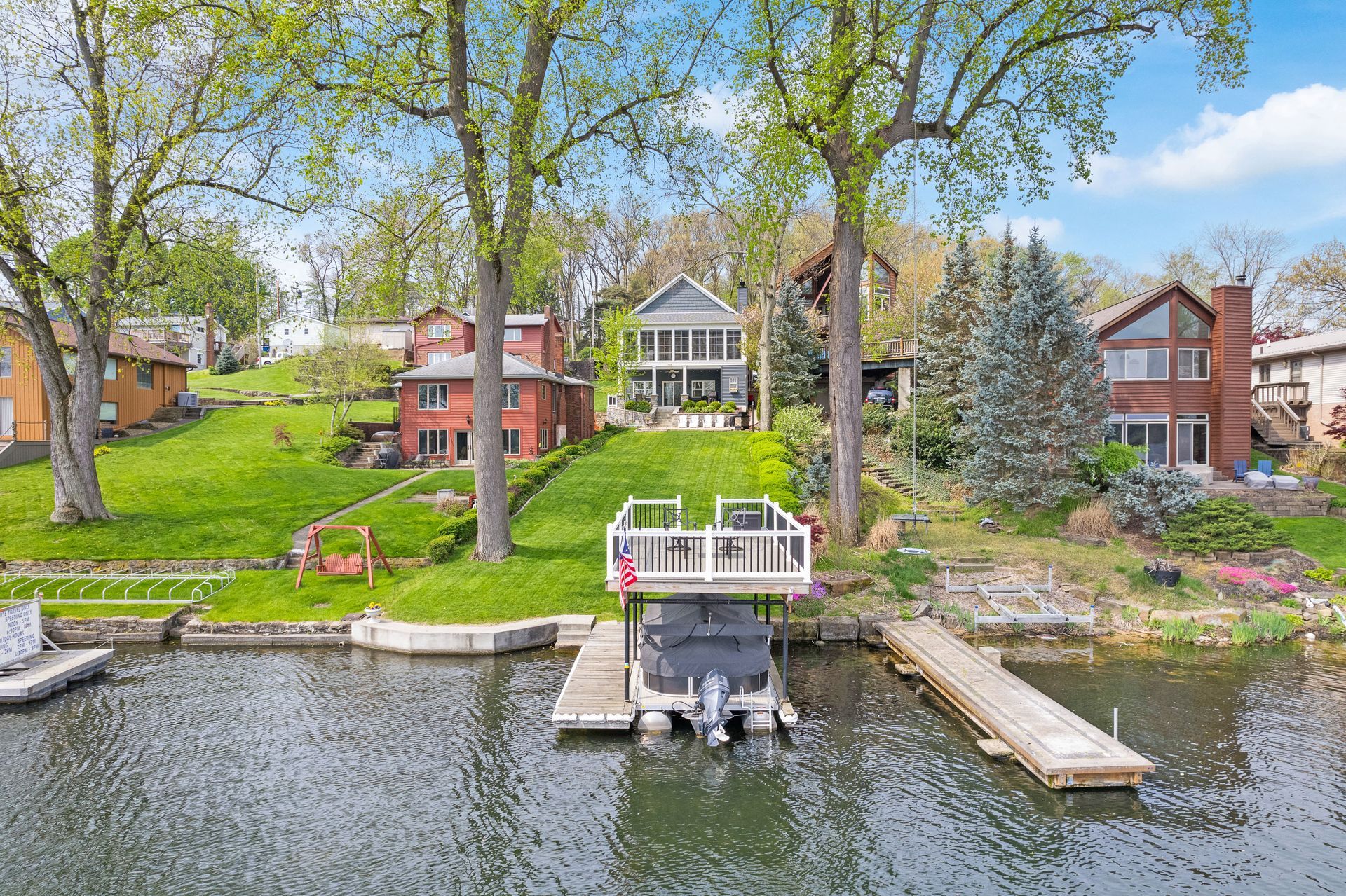 Three houses sit on a grassy, sloped shoreline overlooking a lake with boat docks and a covered boat lift.