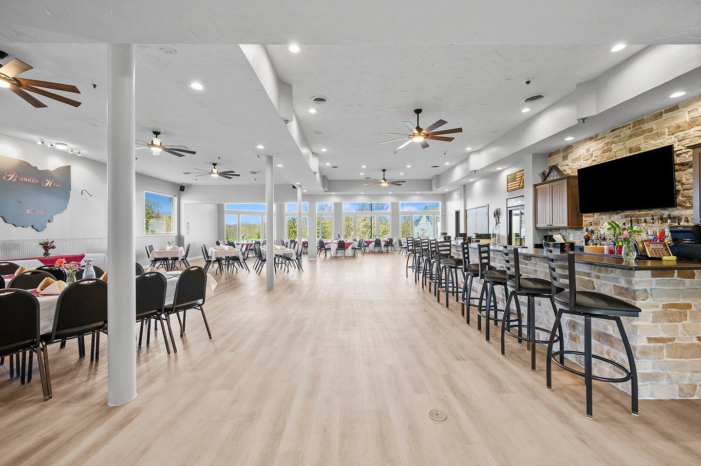 A spacious, modern event hall with tables on the left, light wood floors, and a stone-accented bar with stools on the right.