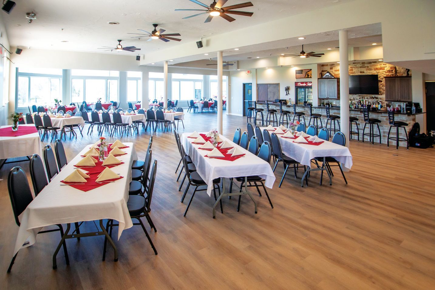 A banquet hall set with long tables covered in white cloths and red runners, facing a bar and windowed seating area.