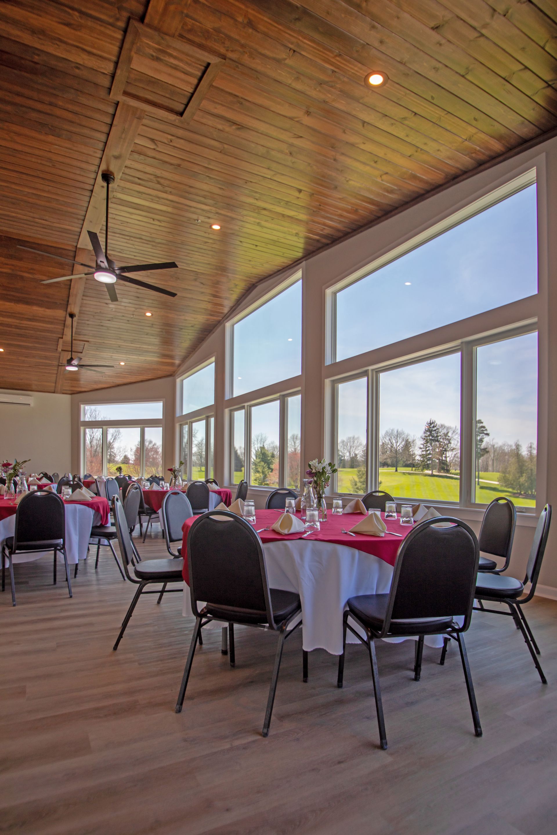 Event space with round tables, red and white linens, and black chairs, set against large windows overlooking a lawn.