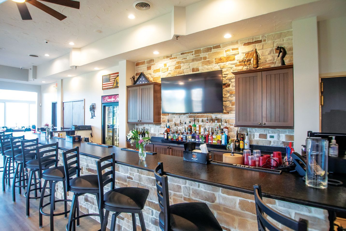 A bar area with stone accents, a television mounted on a wall, brown wooden cabinets, and dark bar stools at the counter.