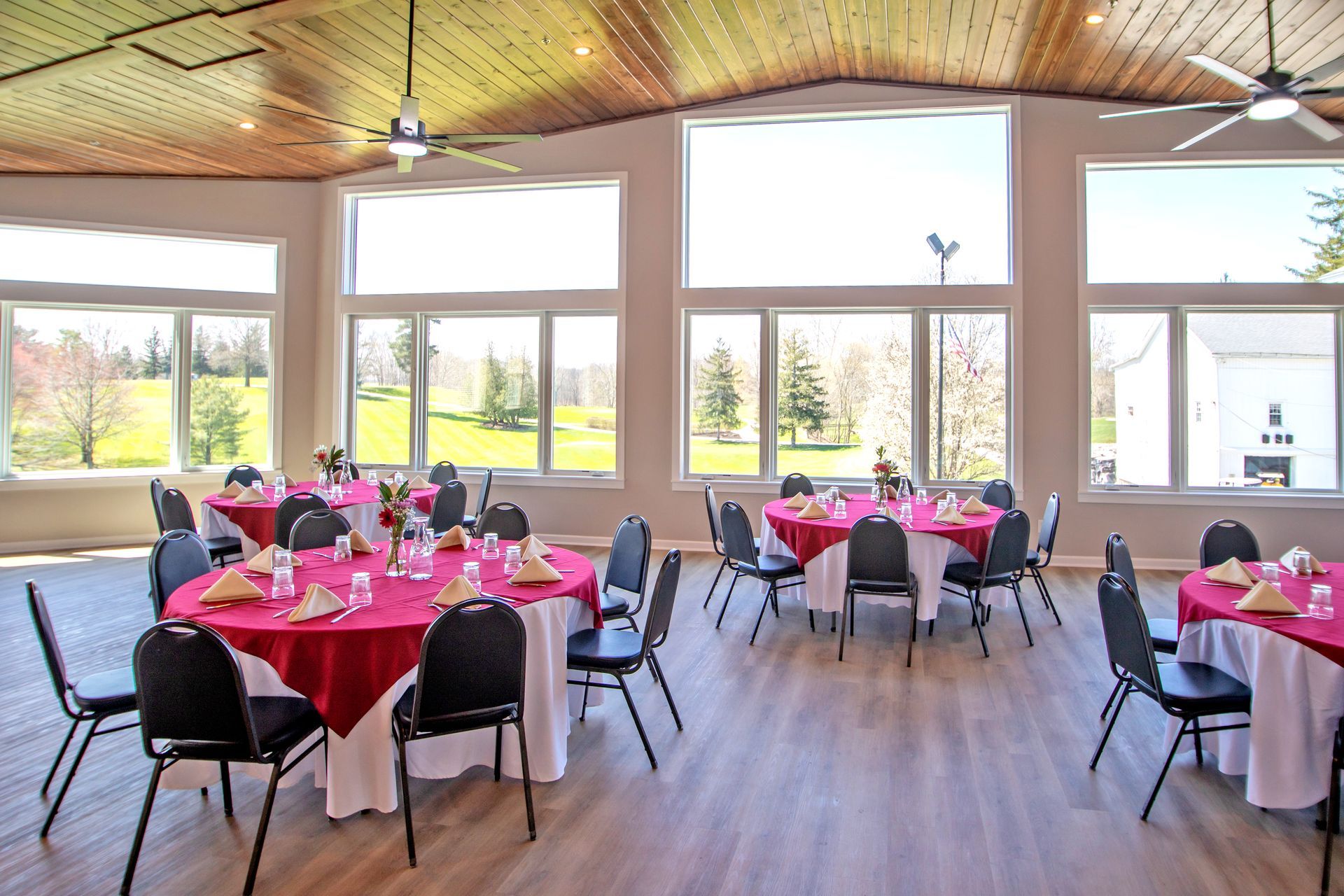 A banquet room with round tables covered in red tablecloths, surrounded by black chairs, set against large window walls.