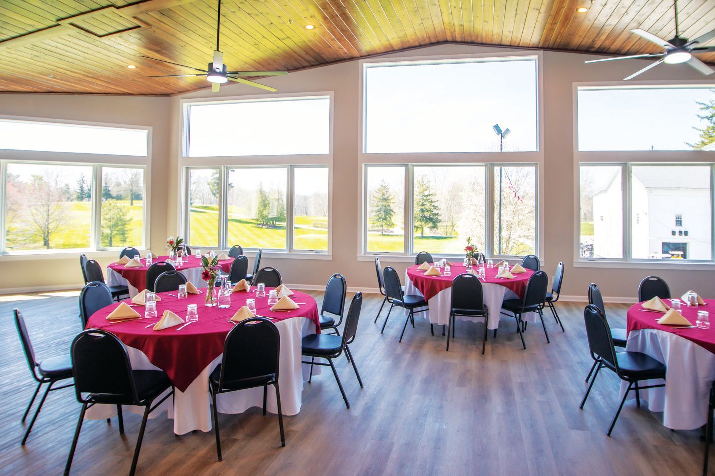 A dining hall with large windows overlooking a field, featuring round tables with burgundy cloths and black chairs.