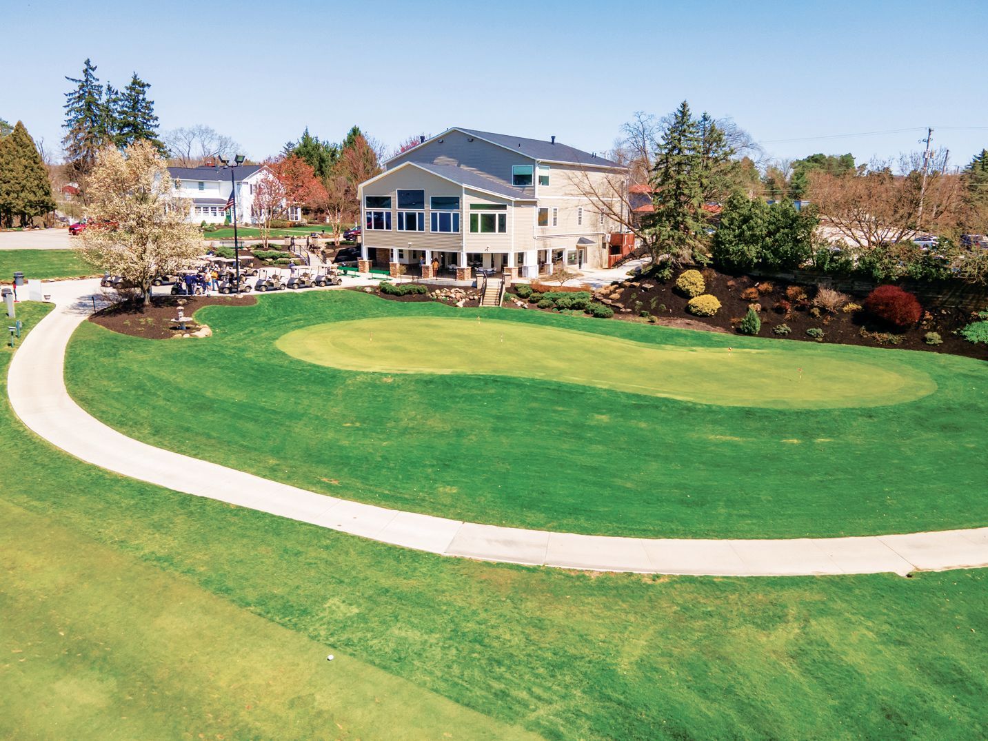 An aerial view of a golf course putting green and path leading toward a large clubhouse on a sunny day.