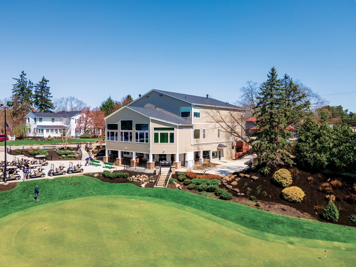 A tan, two-story golf clubhouse building overlooking a green lawn with parked golf carts and trees under a clear blue sky.