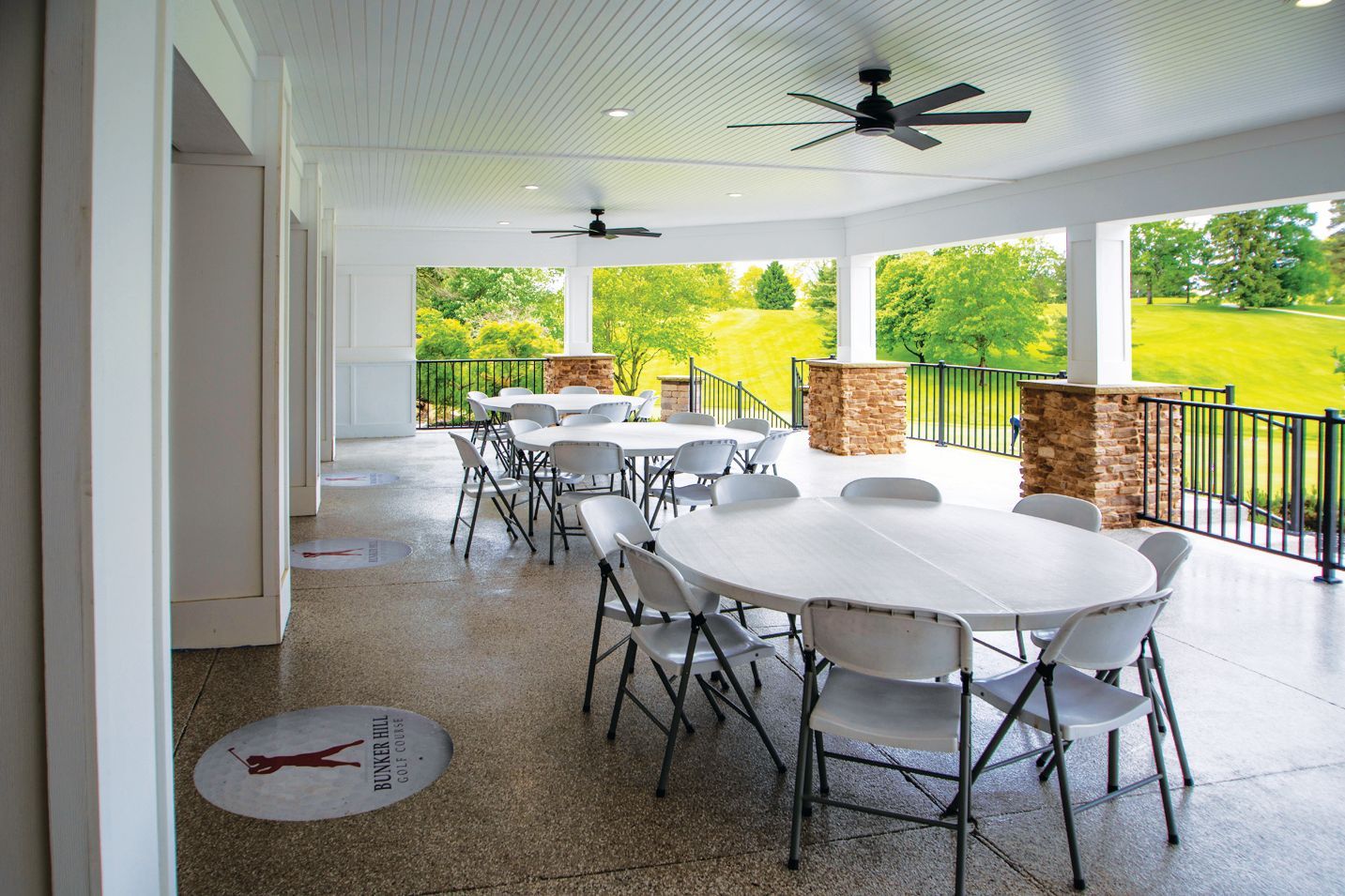 A covered patio with round tables and white folding chairs, overlooking a green landscape with trees and a golf course.