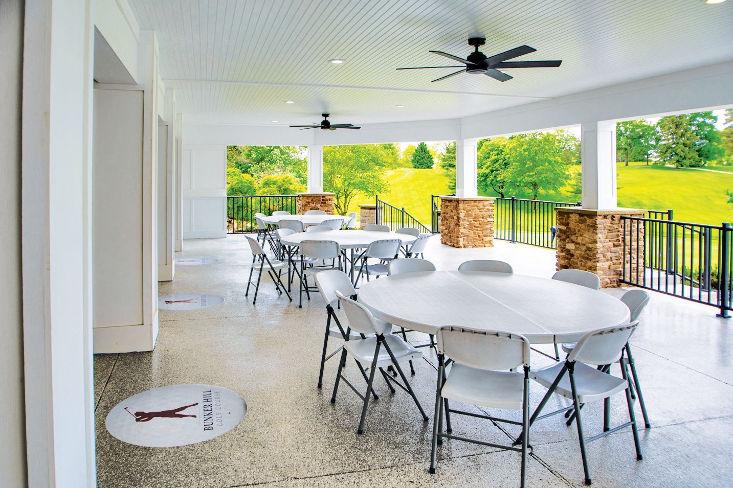 A covered outdoor patio with round white tables and folding chairs, overlooking a grassy hill landscape.