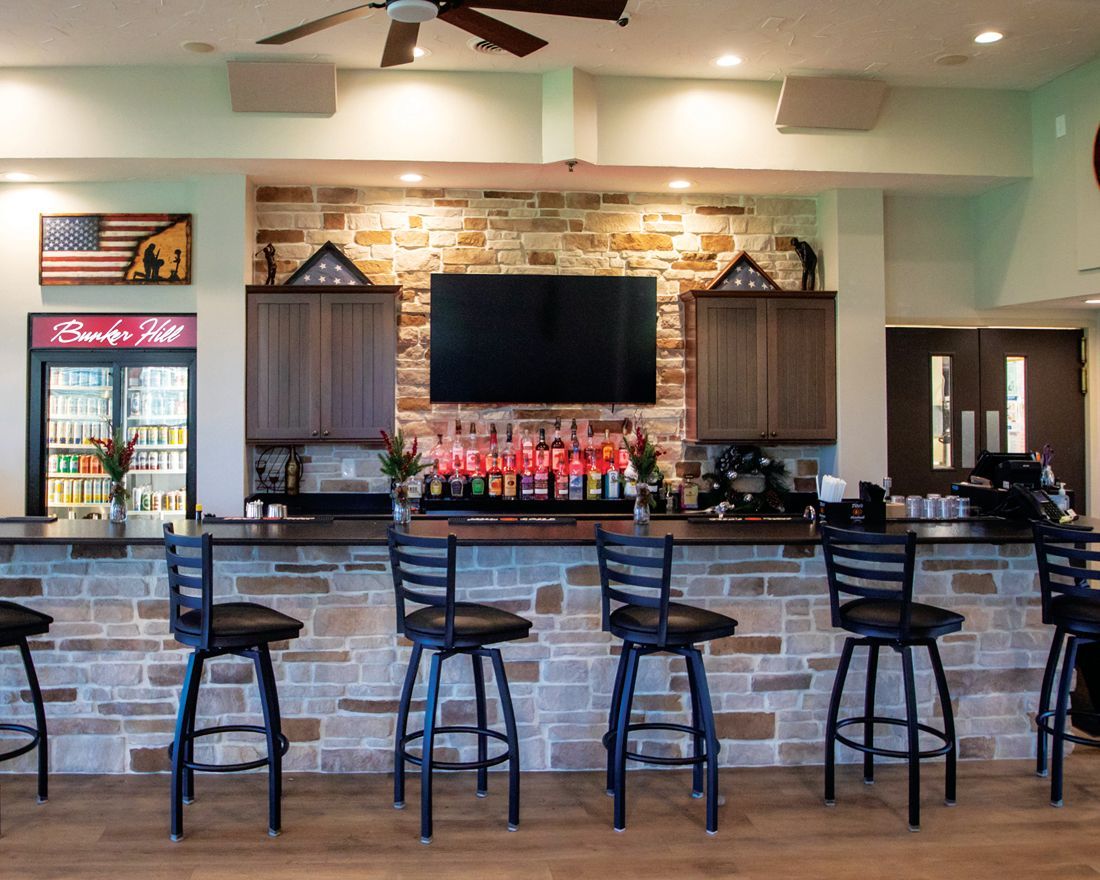 A rustic home bar with a stone wall, flat-screen TV, wooden cabinets, liquor bottles, and five black bar stools.