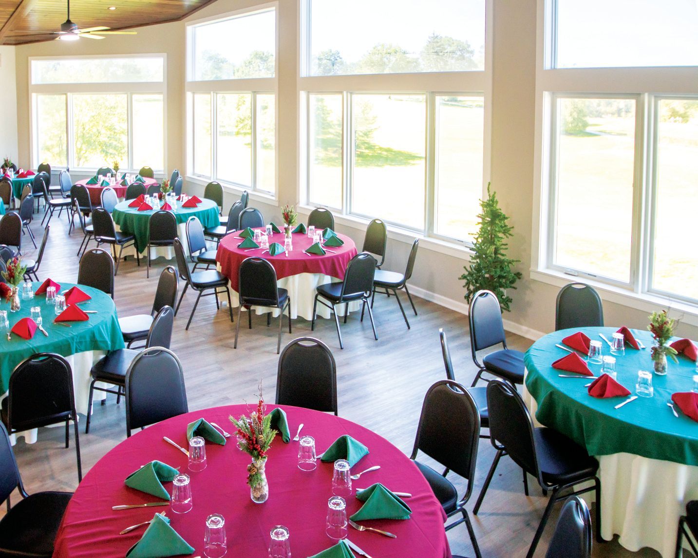A sunlit dining room features round tables with green or maroon tablecloths, black chairs, and tall windows overlooking lawn.