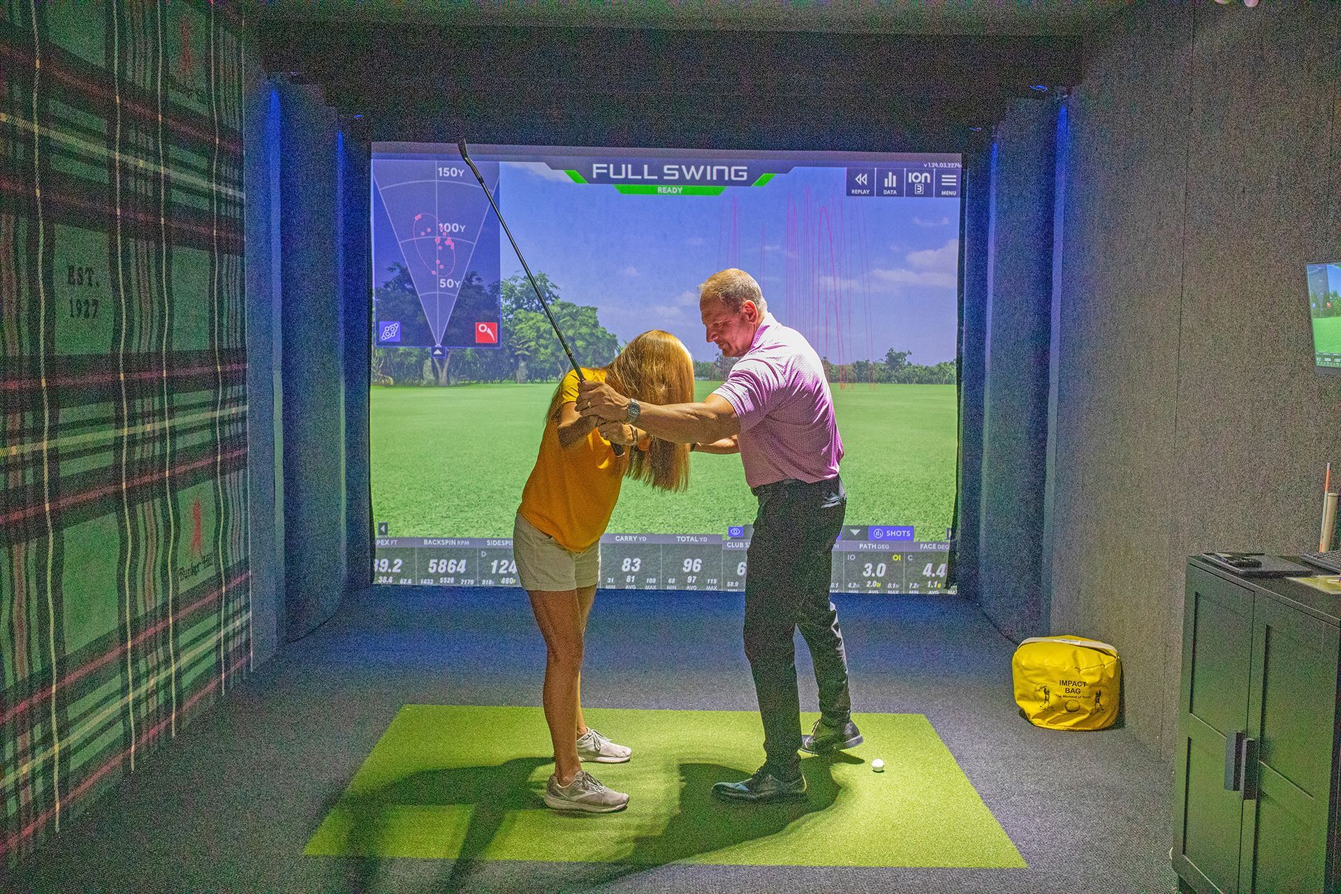 A golf instructor in a purple shirt helps a student in a yellow shirt with their swing at a virtual golf simulator.