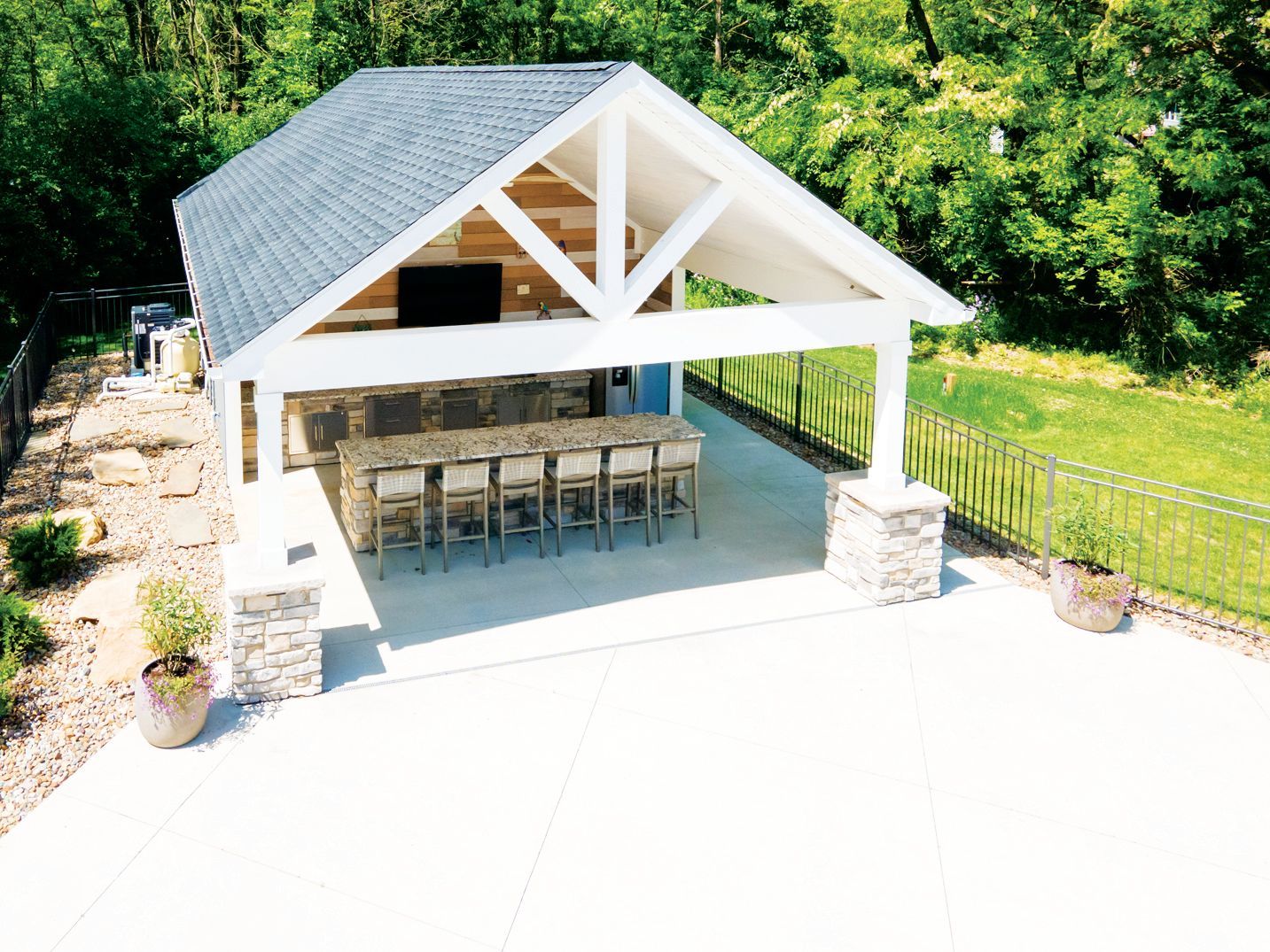 Outdoor pavilion with a bar, stools, and stone columns on a concrete patio, set against a lush, green wooded background.