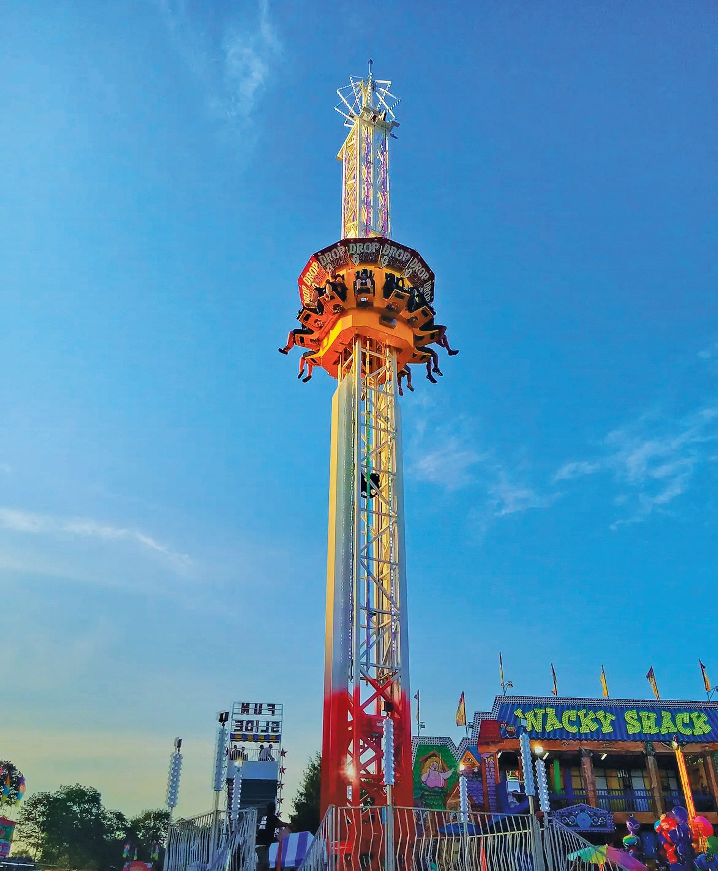 A tall drop-tower amusement ride with riders seated in a circular frame, suspended against a clear blue sky.