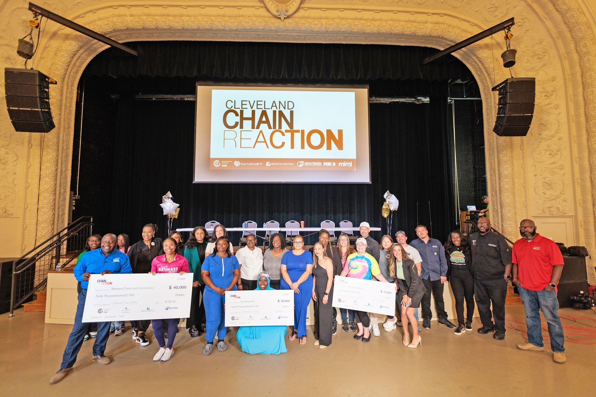 A diverse group of people stands on a stage in front of a Cleveland Chain Reaction sign, holding large ceremonial checks.