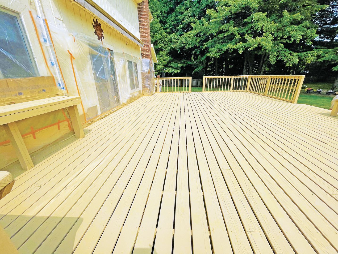 A wide, newly constructed light-yellow wooden deck attached to a house with a brick chimney and green trees in the background.
