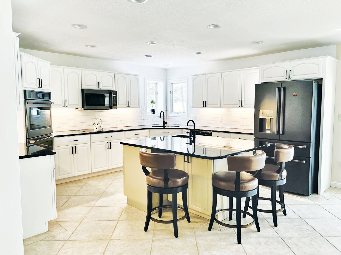 A bright kitchen with white cabinets, black granite countertops, a center island with three stools, and black appliances.