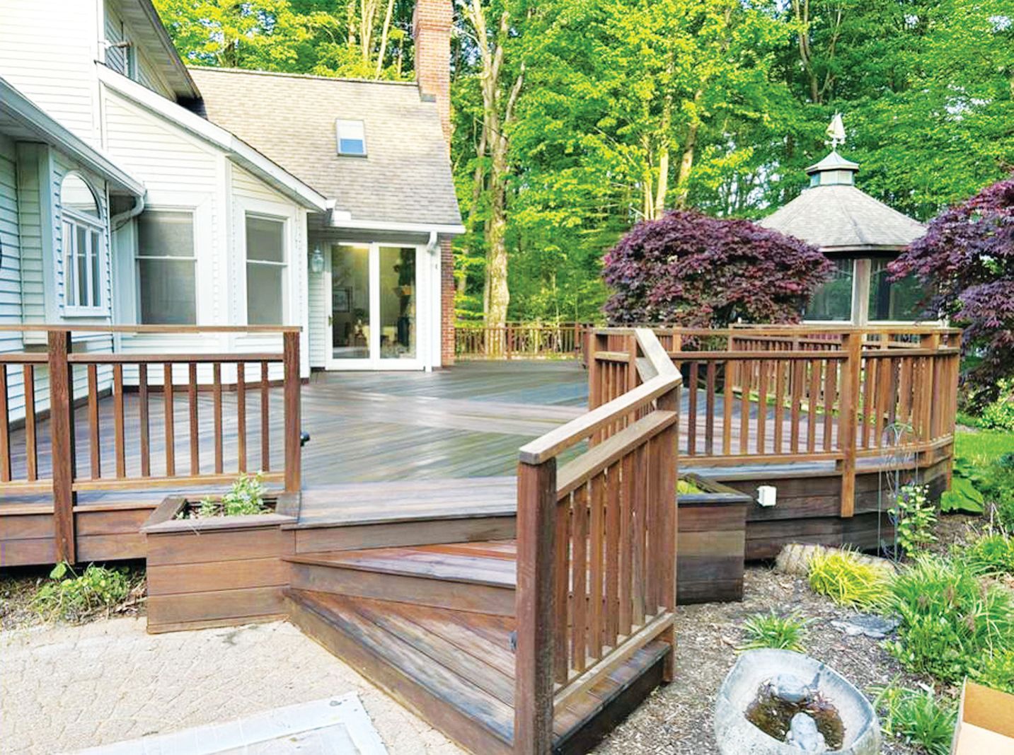 A wooden deck with stairs and railings attached to a house, next to a gazebo in a wooded backyard.