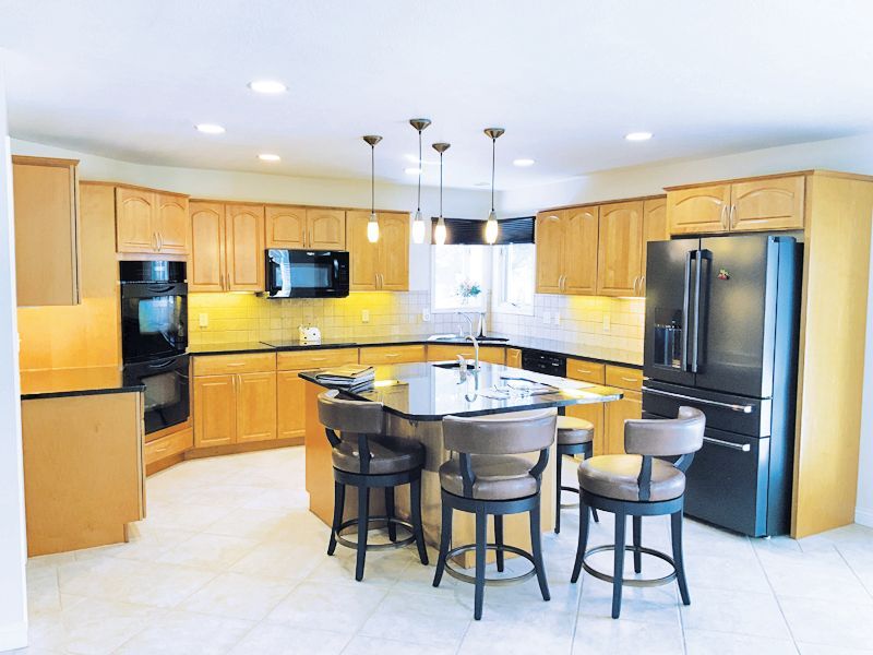 A modern kitchen with wood cabinets, black appliances, a central island with three bar stools, and tiled flooring.