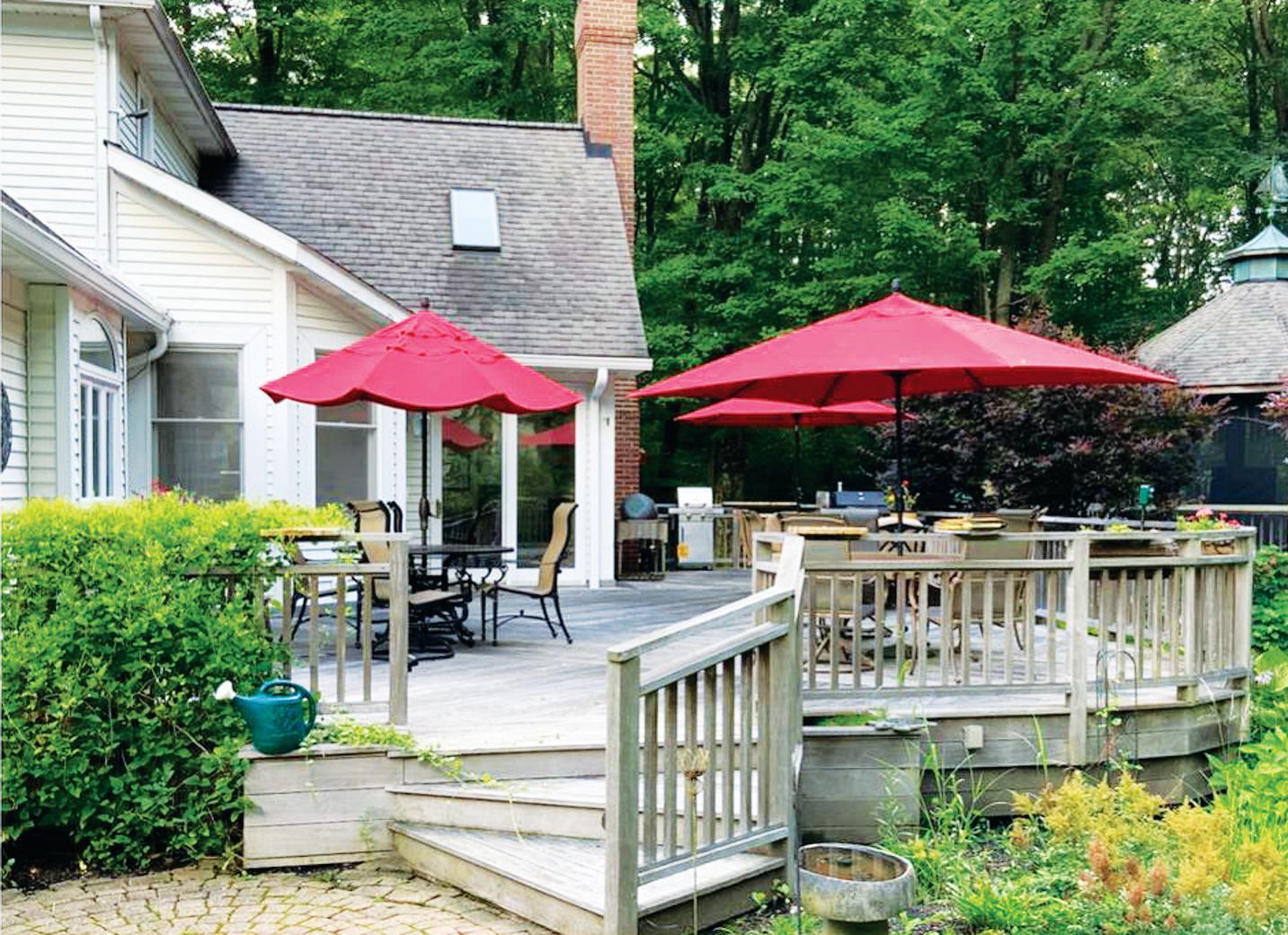 A house with a backyard wooden deck featuring a dining set with red umbrellas, surrounded by lush green trees.